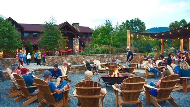 An outdoor seating area with wooden chairs and a central fire pit, surrounded by a stone wall and a building with a red roof. - The Cottage @ Afton Mountain- Close to Wineries! (Afton)