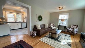 A spacious living area with a brown leather sofa, a coffee table, and patterned rugs. A kitchen is visible in the background with white cabinets and a window. - The Cottage @ Afton Mountain- Close to Wineries! (Afton)