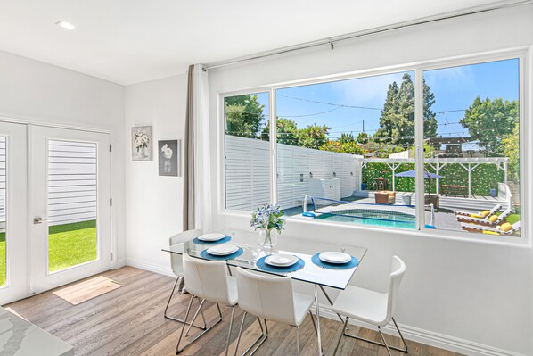 A dining area with a glass table, white chairs, and a view of a backyard with a pool.