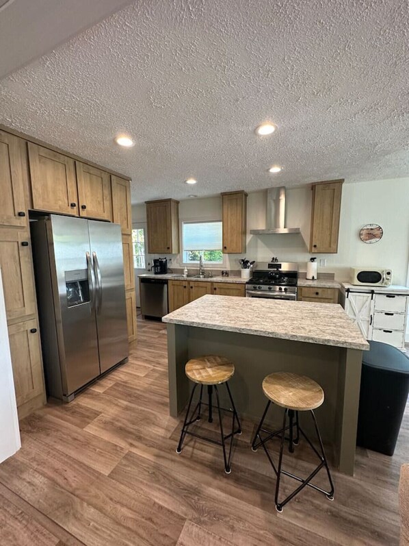 A modern kitchen with wooden cabinets, a stainless steel refrigerator, a central island with stools, and a microwave.