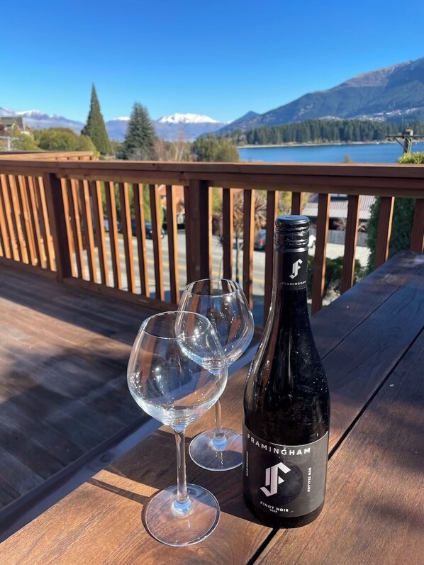 A bottle of Framingham Pinot Noir wine and two empty wine glasses on a wooden deck with a scenic mountain and lake view.