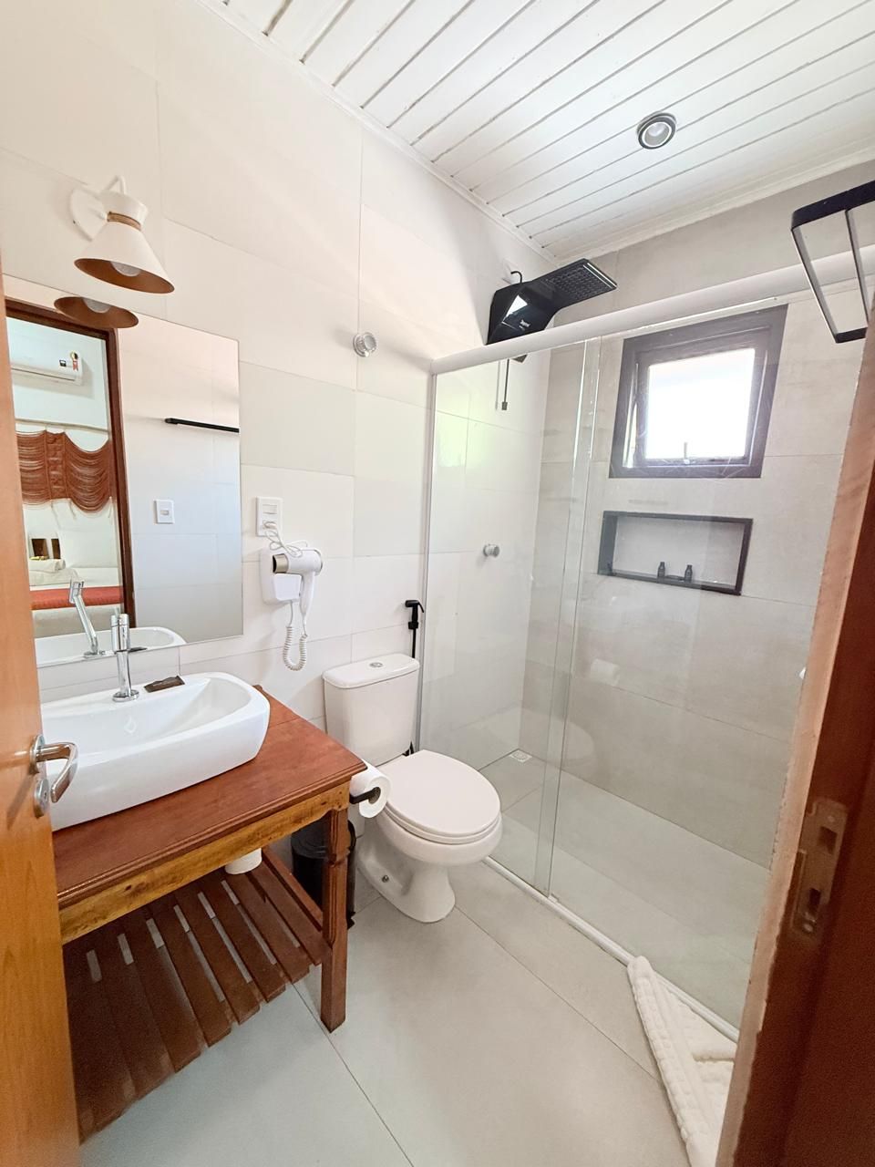 Ein modernes Badezimmer mit einem Holzwaschtisch, einer weißen Toilette, einer Duschkabine aus Glas und einem kleinen Fenster.