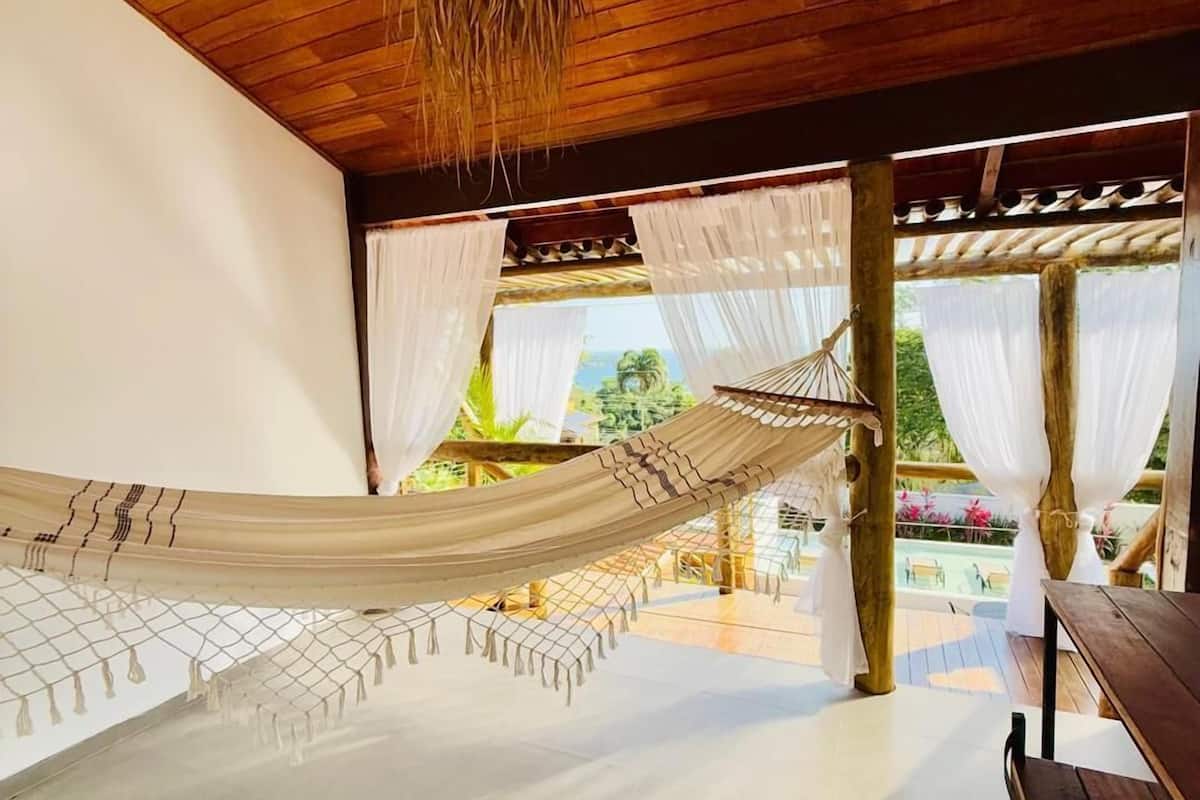 A hammock under a wooden ceiling, a table, and a view of a pool and greenery.