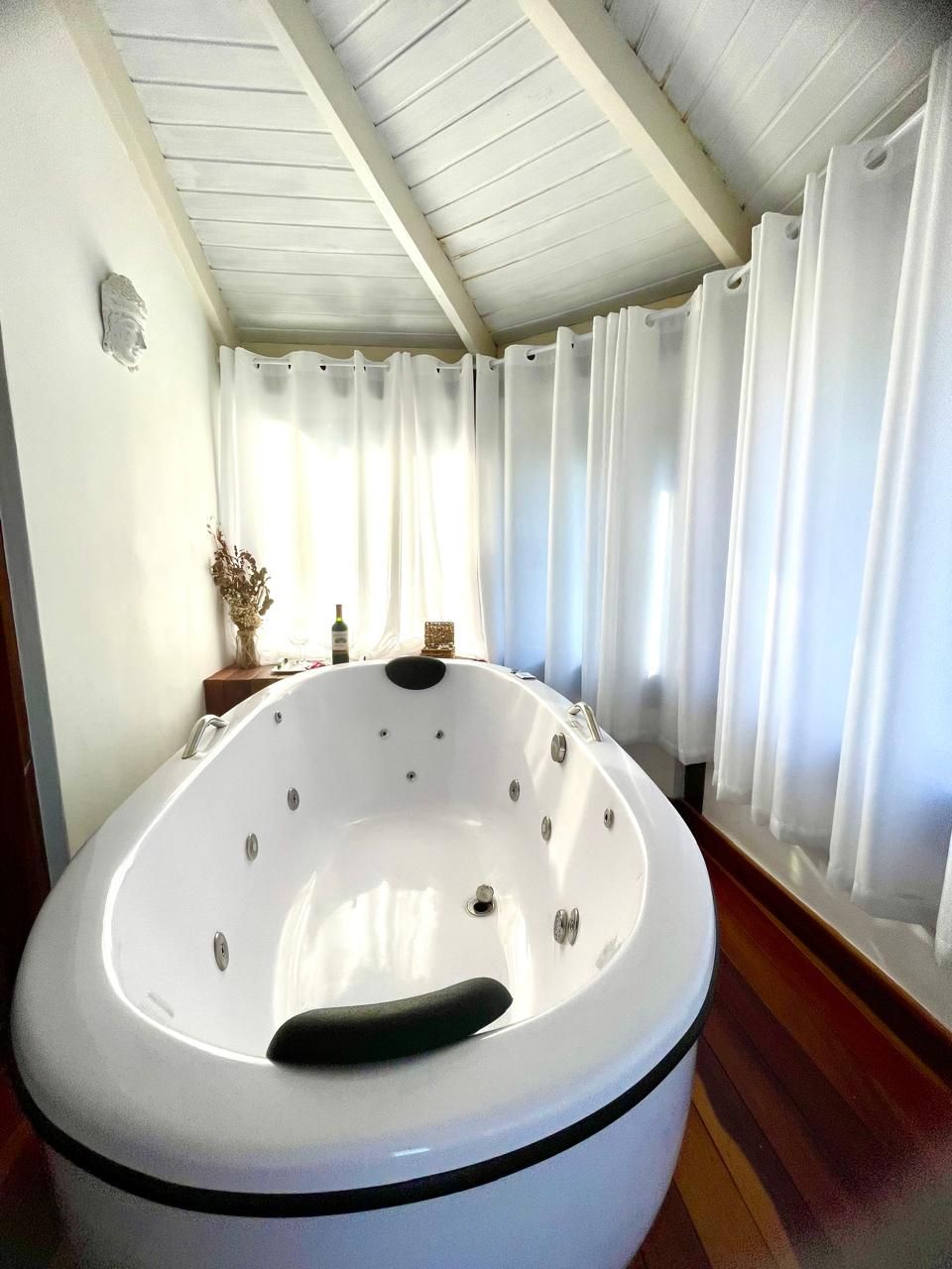 Un grand jacuzzi blanc se trouve dans une pièce dont le sol est en bois et les murs peints en blanc, et des rideaux habillent les fenêtres.