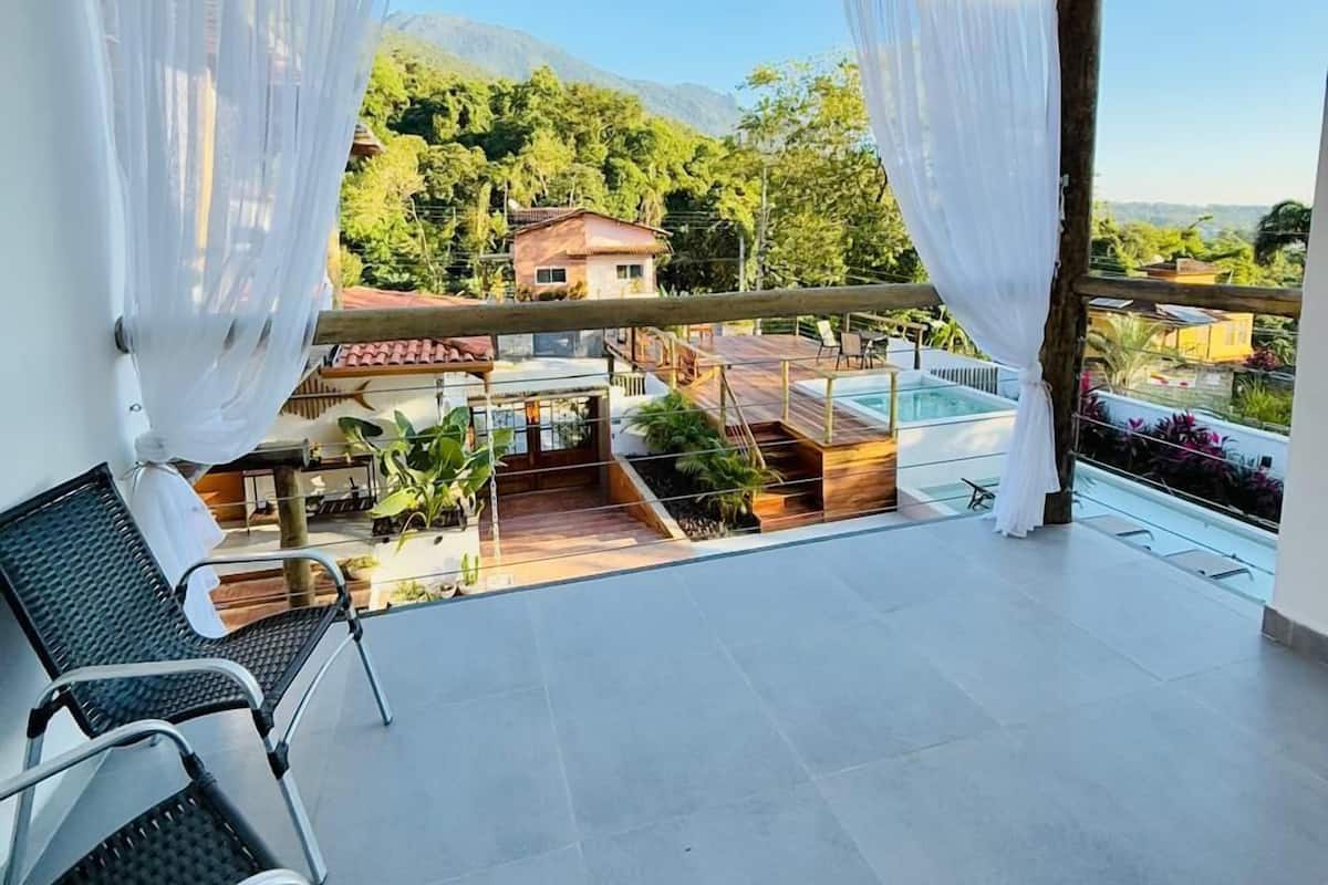 A covered balcony with a pool, white curtains, and a view of a house and mountains.