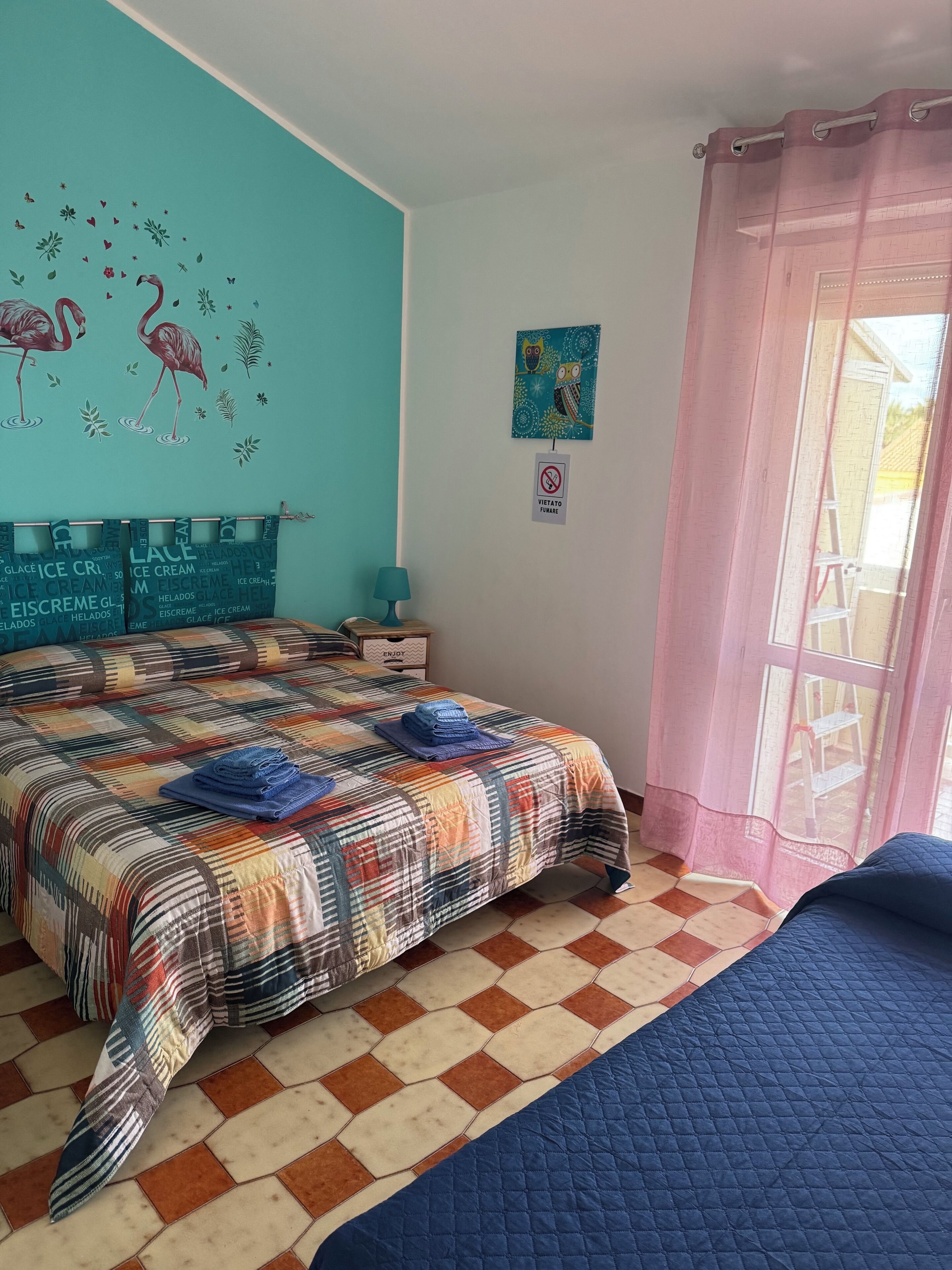 A bedroom with a checkered floor, a bed with a colorful quilt, a wall with a bird mural, and a window with a pink curtain.