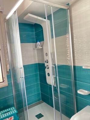 Un baño moderno con ducha envuelta en vidrio, azulejos verde mar y blancos, un pequeño estante con artículos de tocador y un lavamanos blanco.