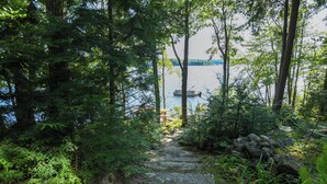 A stone pathway leading to a dock by a lake, surrounded by dense forest. - Designer Lakefront Retreat with Panoramic Sunset Views (Harrison)