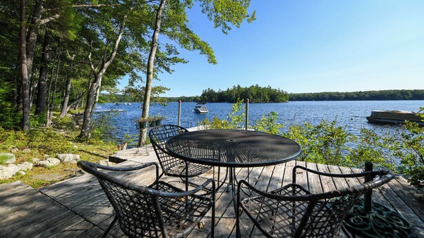 A wooden deck with a round table and chairs overlooking a lake with boats. - Designer Lakefront Retreat with Panoramic Sunset Views (Harrison)