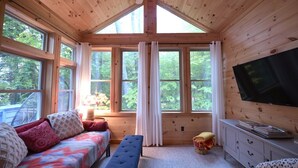 A cozy cabin with a bed, bench, TV, and large windows offering a view of the forest. - Designer Lakefront Retreat with Panoramic Sunset Views (Harrison)