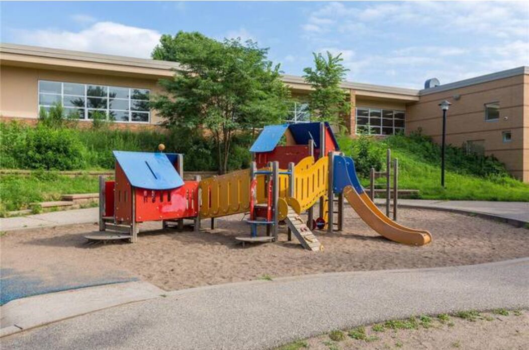 Ein Spielplatz mit Rutsche und Klettergerüsten, umgeben von einem Gebäude mit großen Fenstern und einem befestigten Bereich.