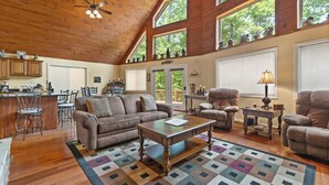 A spacious living area with a wooden ceiling, large windows, a kitchen area, and a cozy seating arrangement. - Large Cabin Located on Deer Water Lake (Rogers)