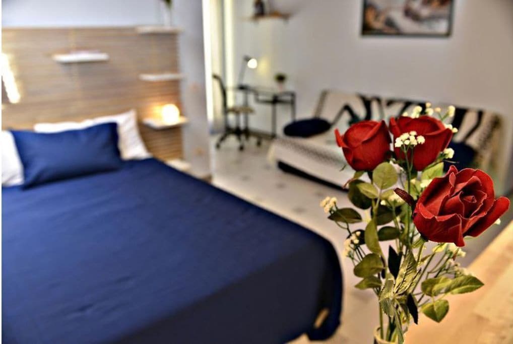 A hotel room with a bed, a nightstand, and a vase with red roses.