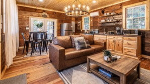 A cozy cabin interior with a wooden ceiling, a chandelier, a leather sofa, a dining area, and a kitchen with wooden cabinets and appliances.