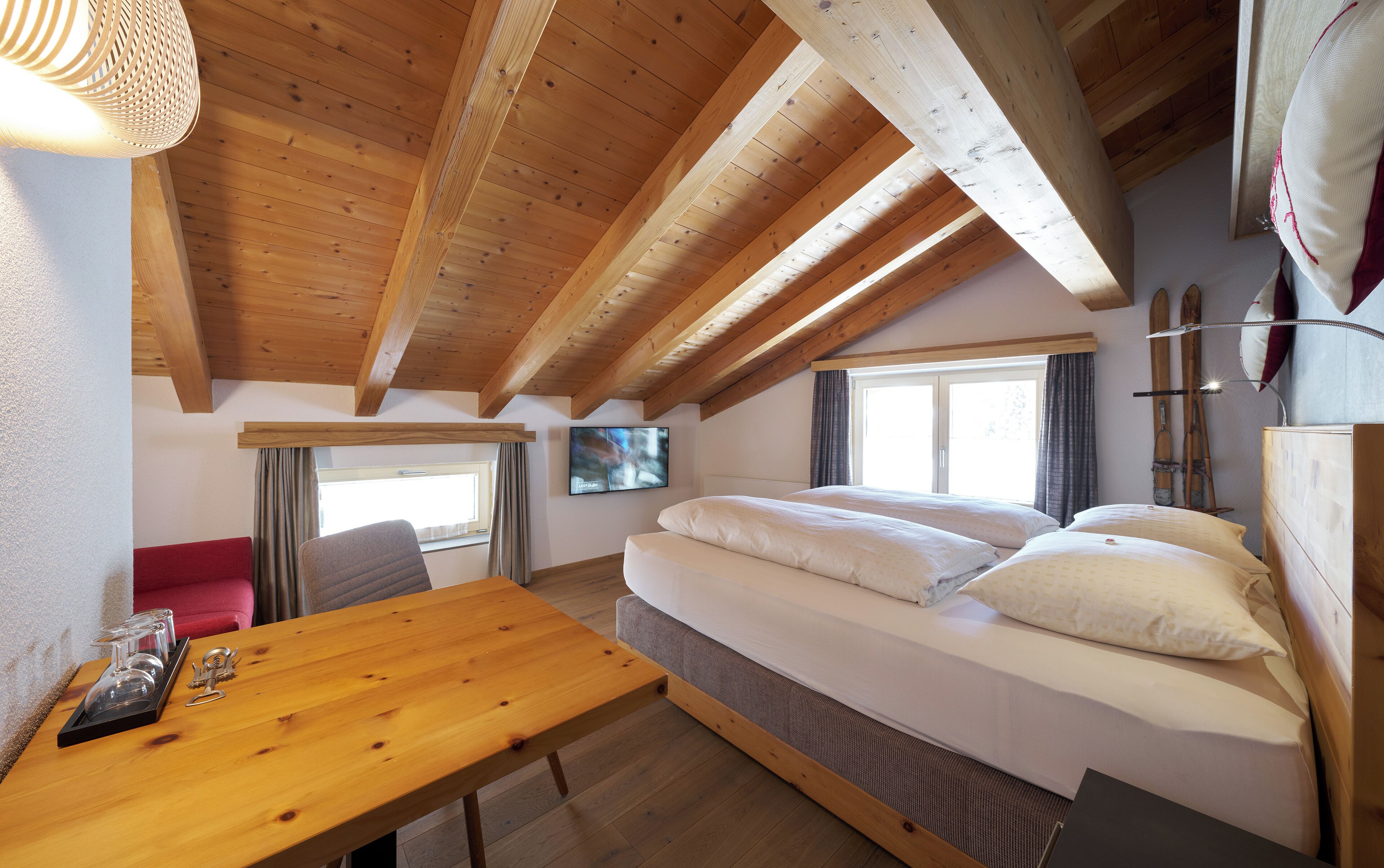 Una habitación acogedora con techo de madera, una cama con ropa blanca, una mesa de madera con un recipiente de vidrio y una silla roja.