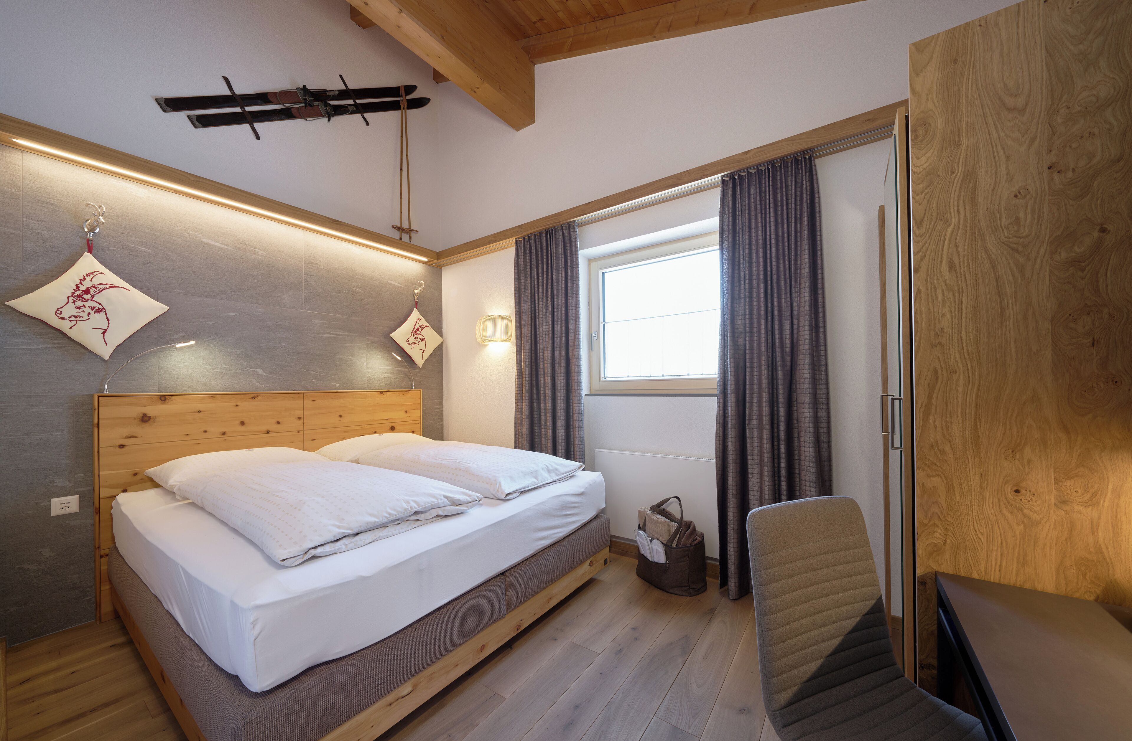 A bedroom with a wooden bed, a chair, a window with curtains, and a ski rack on the wall.