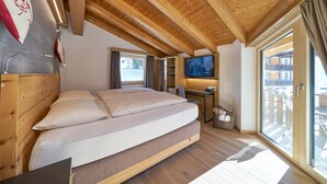 A bedroom with a bed, wooden ceiling, a TV, and a window with a view of buildings.