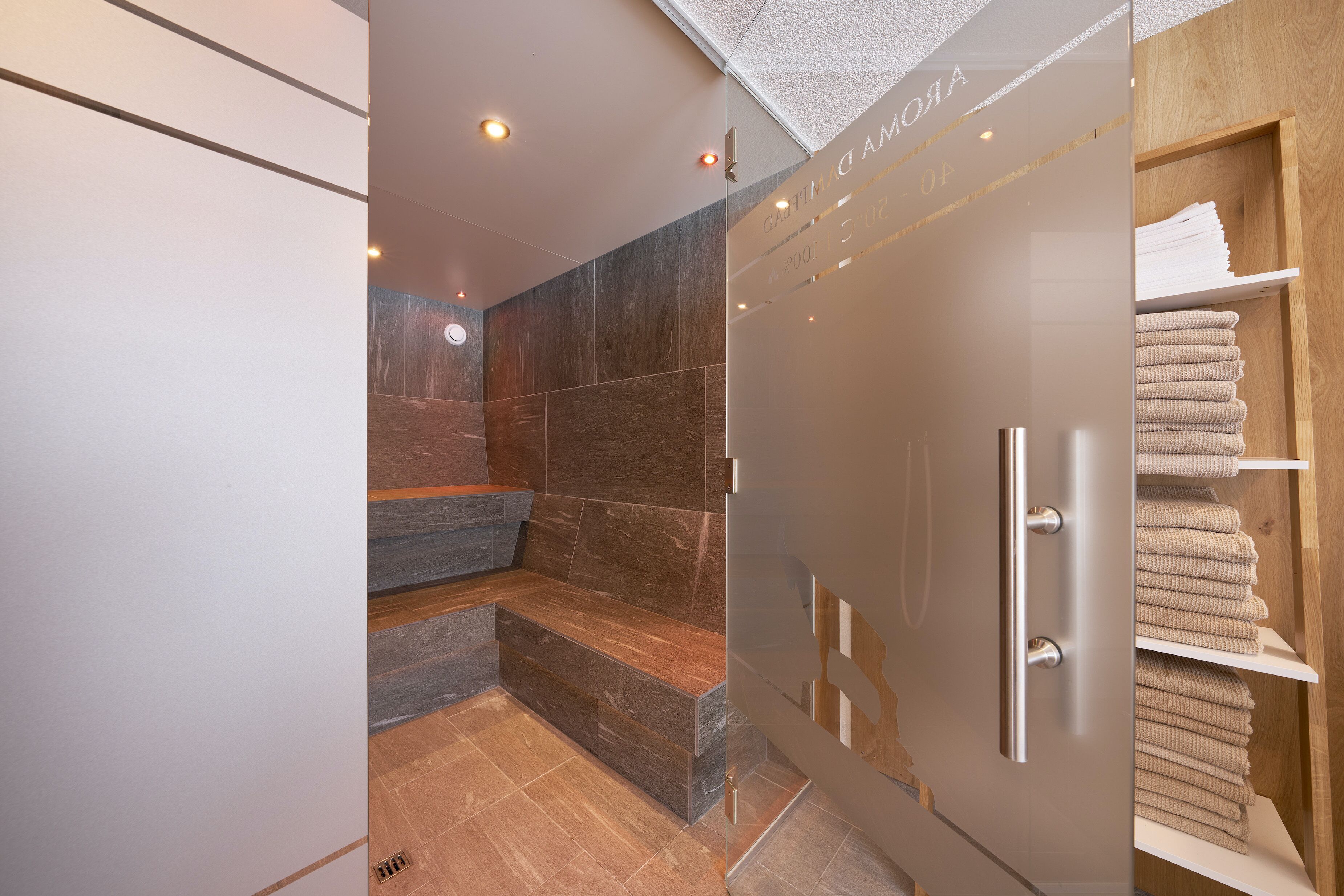 A modern bathroom with a glass shower enclosure, a marble-tiled wall, and a wooden shelf with towels.