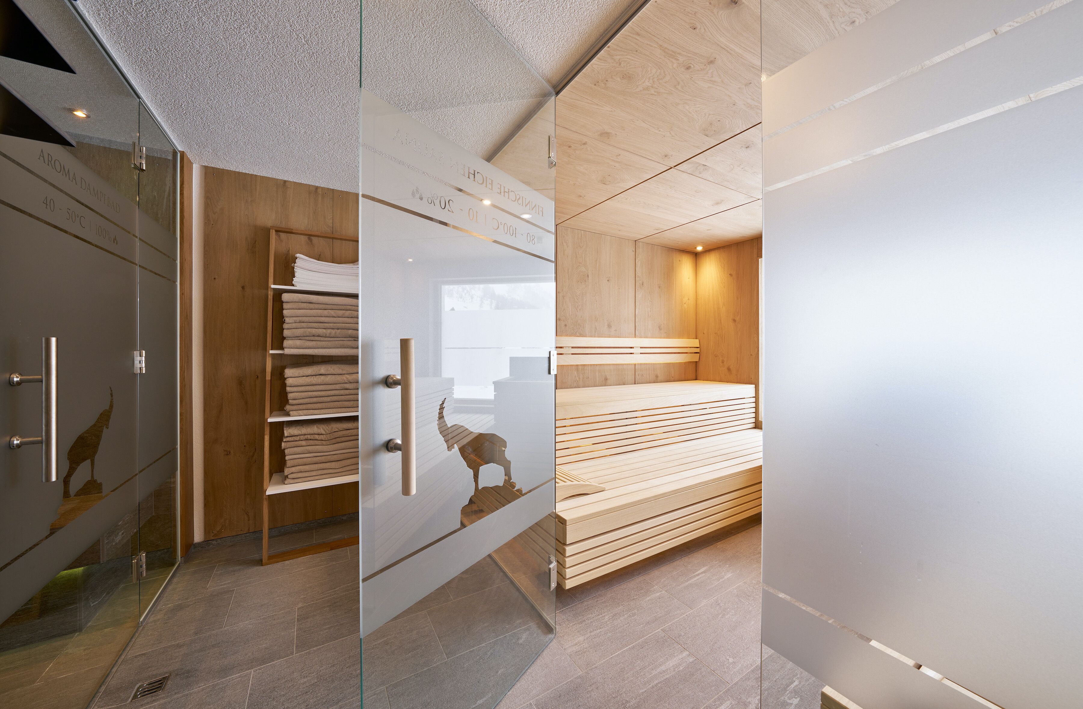 A modern bathroom with a glass shower enclosure, wooden shelving with towels, and a wooden bench.