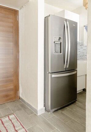 A stainless steel refrigerator in a kitchen with a wooden door and tiled flooring.