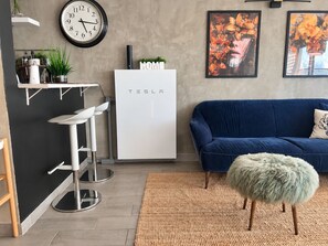 A modern living room with a blue sofa, a white Tesla refrigerator, a round wall clock, and a bar area with stools.