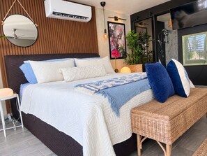 A modern bedroom with a large bed, a bedside table, a wall-mounted air conditioner, a hanging mirror, and a window with a view.