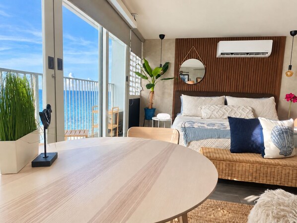 A modern bedroom with a large bed, a round dining table, and a balcony with a view of the sea.