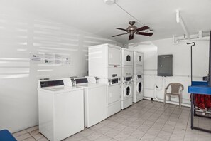 A laundry room with stacked washers and dryers, a ceiling fan, a chair, and a blue table with a red cloth.