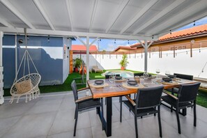 An outdoor patio with a dining table set for four, surrounded by chairs, a hanging chair, and a small garden area.