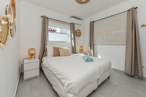 A bedroom with a bed, two bedside tables, a window with curtains, and a wall-mounted air conditioner.