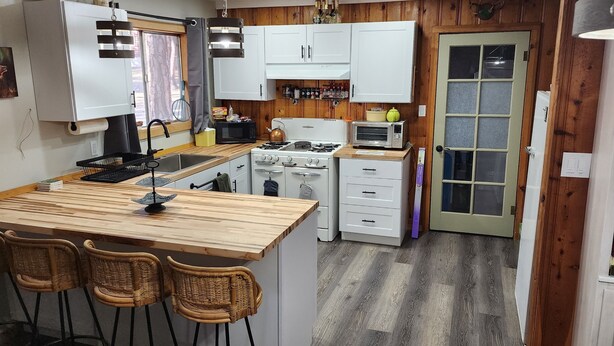A compact kitchen with a central island, white cabinets, and a stove. There are wicker chairs around the island and a small dining table. - Alpine Dog Friendly Cabin! (Alpine)