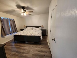 A bedroom with a bed, a nightstand, a lamp, a ceiling fan, and a window with curtains. - The Lynn Inn (Long Beach)