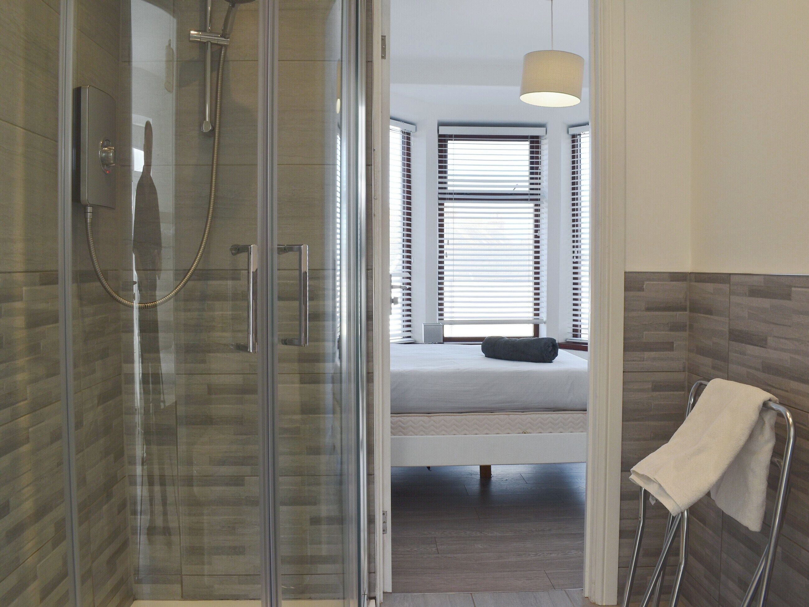 Un baño moderno con cabina de ducha de vidrio, una cama visible con colchón blanco y un perchero con toalla blanca.