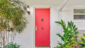 Eine rote Tür mit der Nummer 2, eine Fußmatte und verschiedene Pflanzen rund um den Eingang.