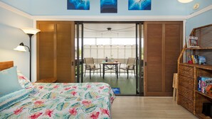 A bedroom with a bed, a nightstand, a lamp, a bookshelf, and a sliding glass door leading to a balcony with a table and chairs. - Happy Honu Oasis, Surf shack with pool and A/C (Hilo)
