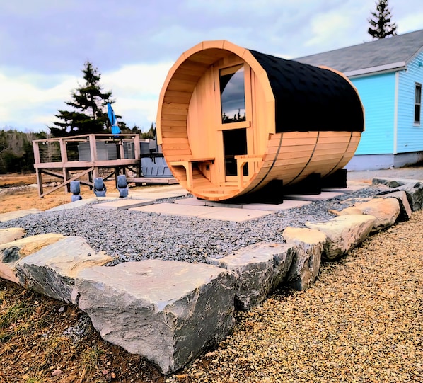 Une structure cylindrique en bois, dotée d’une porte, entourée d’un chemin de gravier et bordée de pierres, avec un bâtiment bleu en arrière-plan.