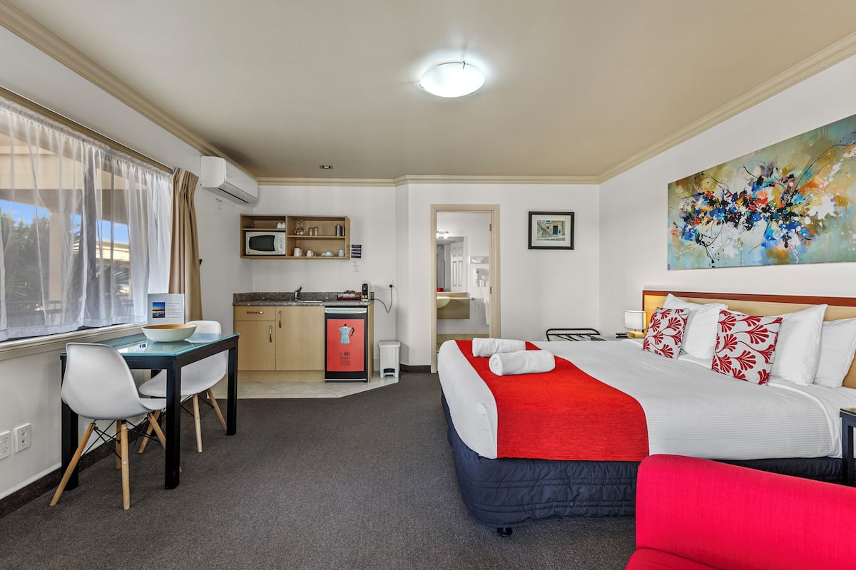 A hotel room with a bed, a desk, a chair, a microwave, a sink, and a painting on the wall.