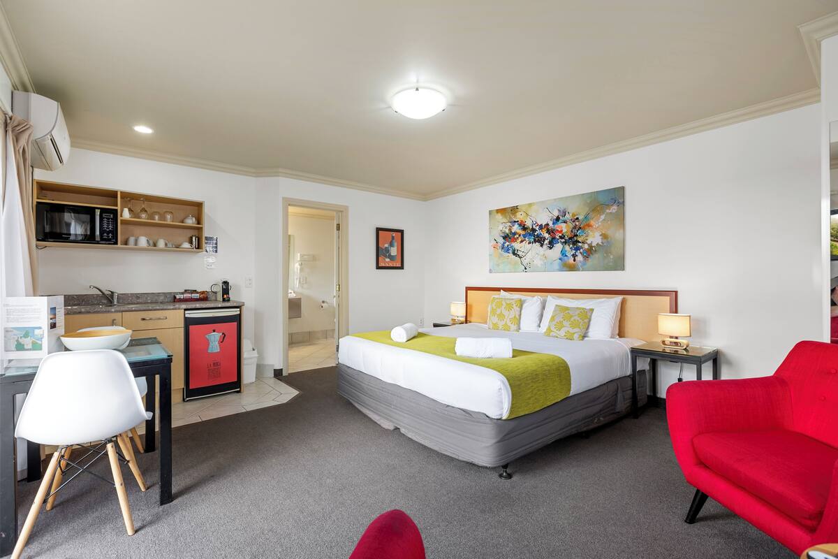A hotel room with a bed, a desk with a computer, a red chair, a red couch, a microwave, a sink, and a painting on the wall.