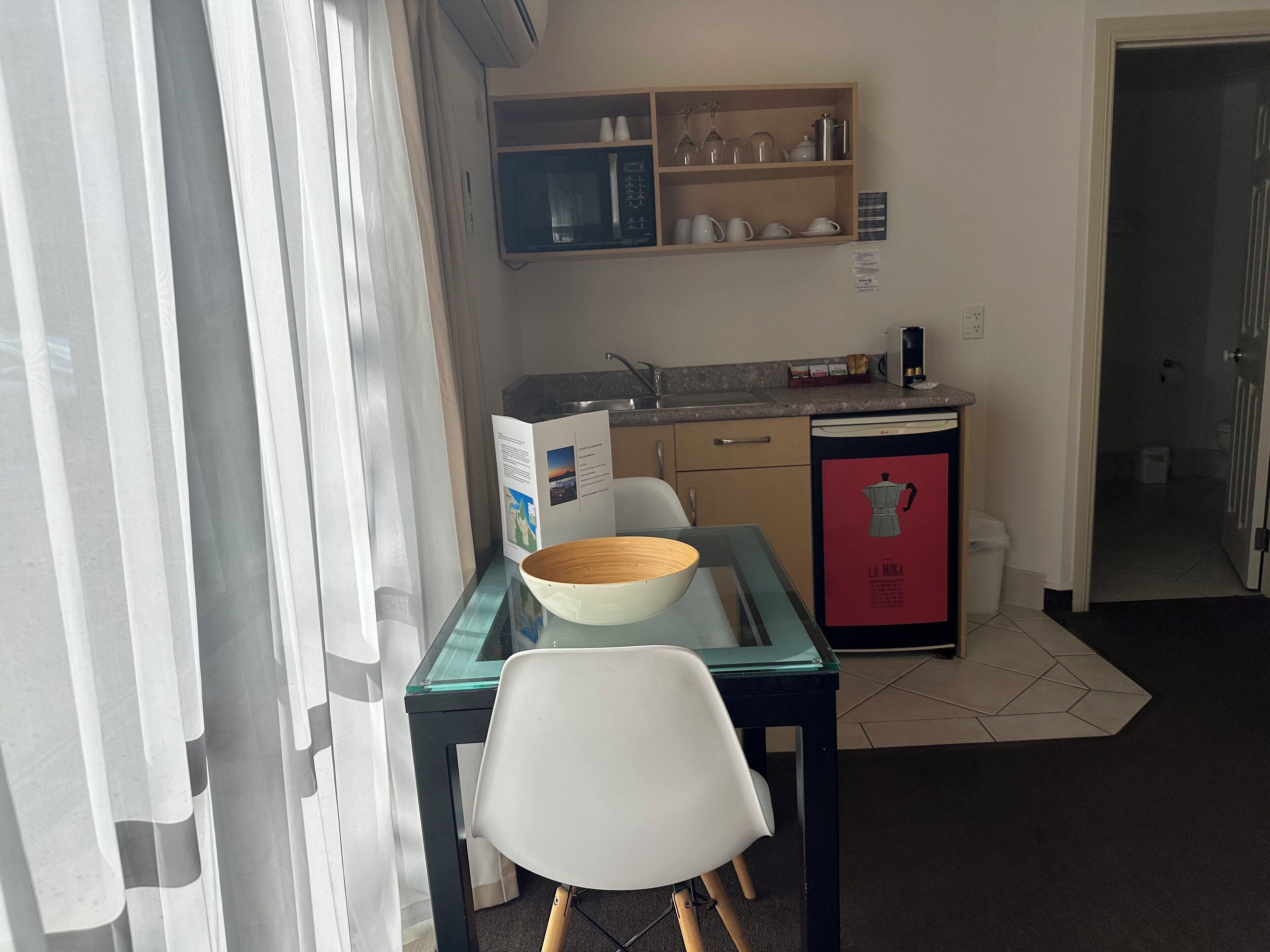 A compact kitchen with a microwave, sink, and a red refrigerator. There's a dining table with a glass top and a white chair.