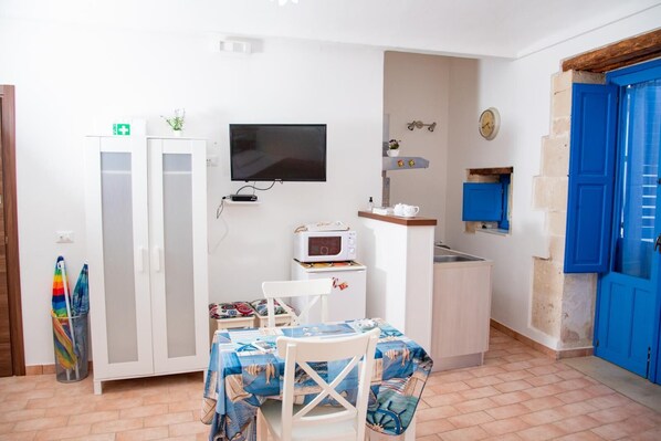A compact room with a kitchenette, a dining area with a table and chairs, a television, and a blue door.