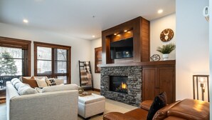 A living room with a stone fireplace, a television mounted above it, and a clock on the wall. - Tremblant Prestige - Étoile 1510-4 (Mont-Tremblant)