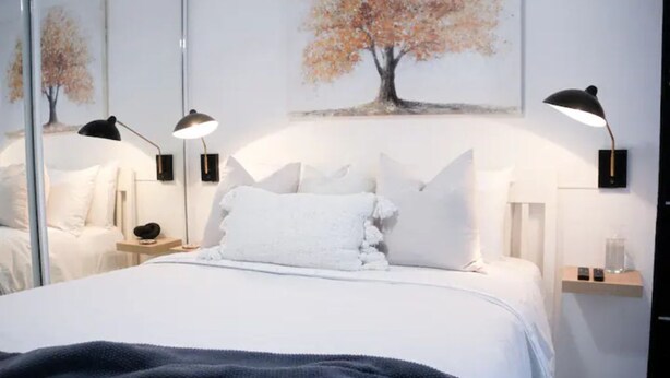 Een keurig opgemaakt bed met witte lakens, twee bedlampen en een schilderij van een boom aan de muur.