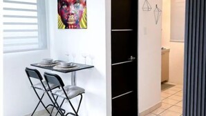 Een compacte leefruimte met een eethoek, bestaande uit een tafel en stoelen, een zwart wandrek, een kleurrijk kunstwerk aan de muur en een klein keukenhoekje op de achtergrond.
