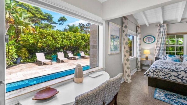 A bedroom with a large window, a pool, and a view of tropical vegetation. - Perfect Urban Escape: Your Personal Oasis with a Zen Den and Lush Gardens! (Delray Beach)