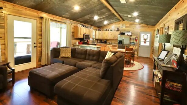 A cozy cabin interior with a sectional sofa, wooden floors, a kitchenette, and a ceiling fan. - Old Mill Cabin #1 - on the Little MO River, large yard, close to Diamond Mine! (Murfreesboro)