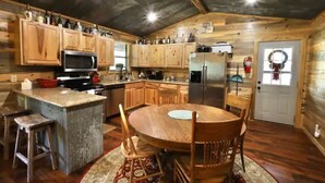 A rustic kitchen with wooden cabinets, a central island, and a dining table. - Old Mill Cabin #1 - on the Little MO River, large yard, close to Diamond Mine! (Murfreesboro)