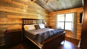 A wooden cabin bedroom with a bed, nightstand, and a window with curtains. - Old Mill Cabin #1 - on the Little MO River, large yard, close to Diamond Mine! (Murfreesboro)