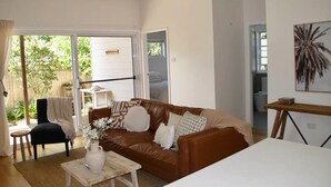 A living room with a brown leather sofa, a white coffee table, and a dining table with chairs. There is a sliding glass door leading to a patio area with a small table and chairs. - WERRI North.
 (Werri Beach)