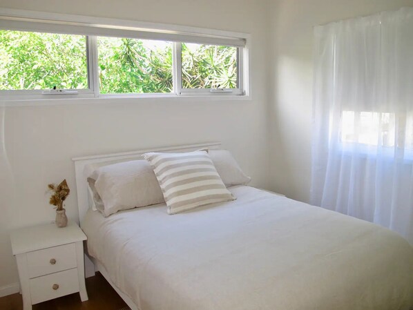A bedroom with a bed, a nightstand, a window with curtains, and a view of greenery outside. - WERRI North.
(Werri Beach)