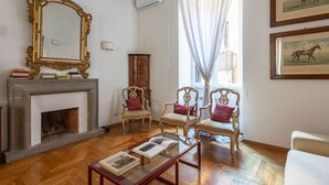 Una sala de estar con chimenea, tres sillones, una mesa de centro con libros, un espejo grande y cuadros en la pared.
